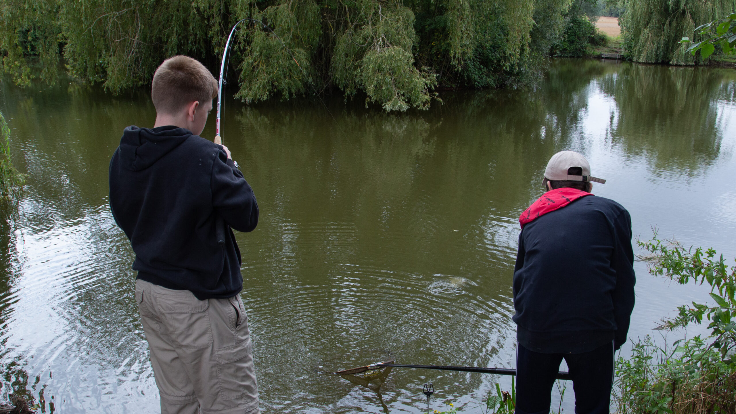 Two boys fish in front of a pond surrounded by green trees