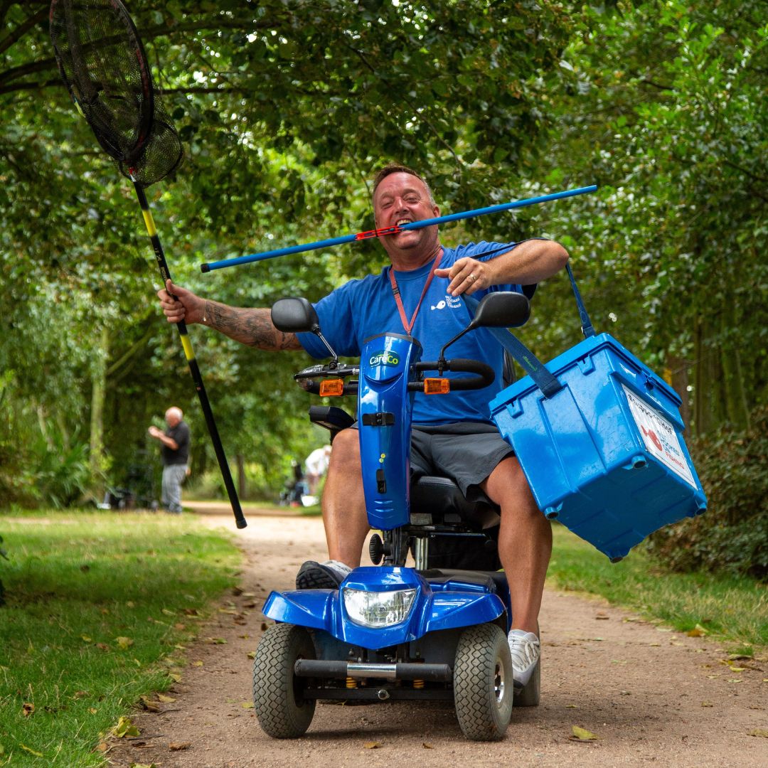 Carl from Get Hooked on Fishing is riding a mobility scooter holding a net, rod and tackle box