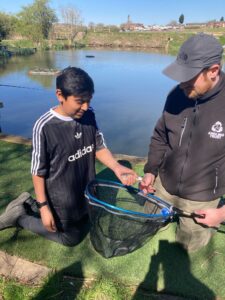 A boy proudly holds his first catch from the freshwater lake at Starmount Fishery with Coach Chris with a backdrop of the lake